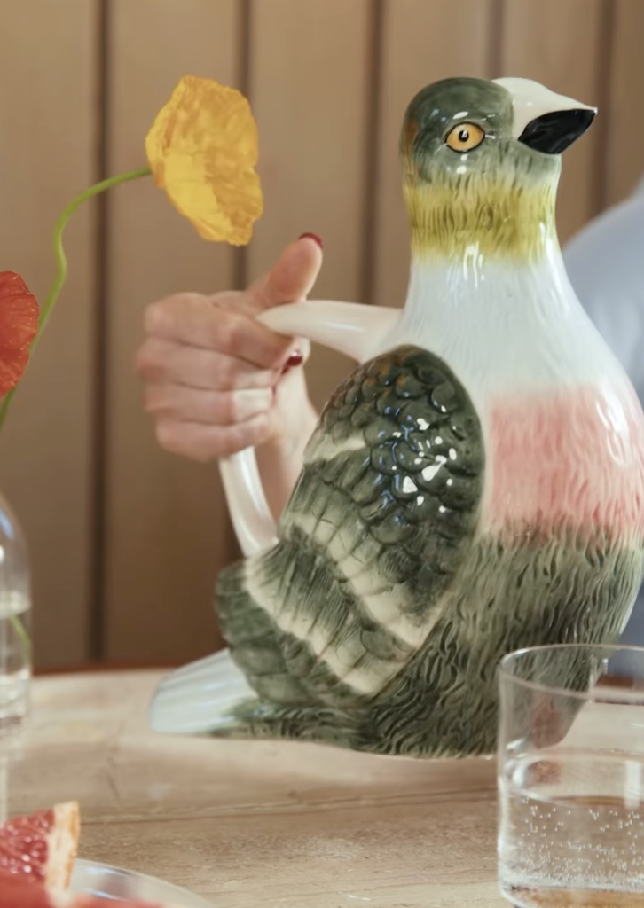 Main tenant une carafe en céramique en forme d'oiseau de la collection maison Emma Chamberlain x West Elm, stylisée sur une table en bois avec des verres et des fleurs