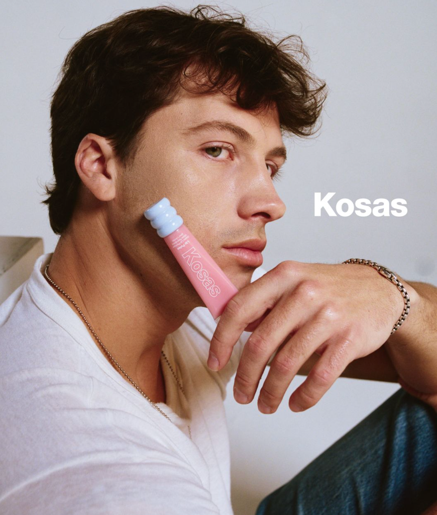Rob Rausch tenant un produit lèvres Kosas rose contre sa joue dans une image de campagne en gros plan pour le partenariat de marque Kosas