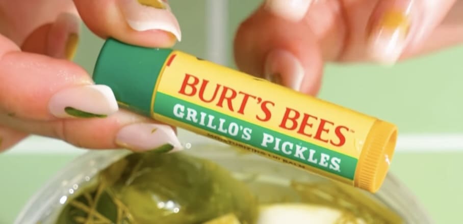 A hand holding a Burt's Bees lip balm tube labelled "Grillo's Pickles" in yellow and green packaging, held over a bowl of pickles.
