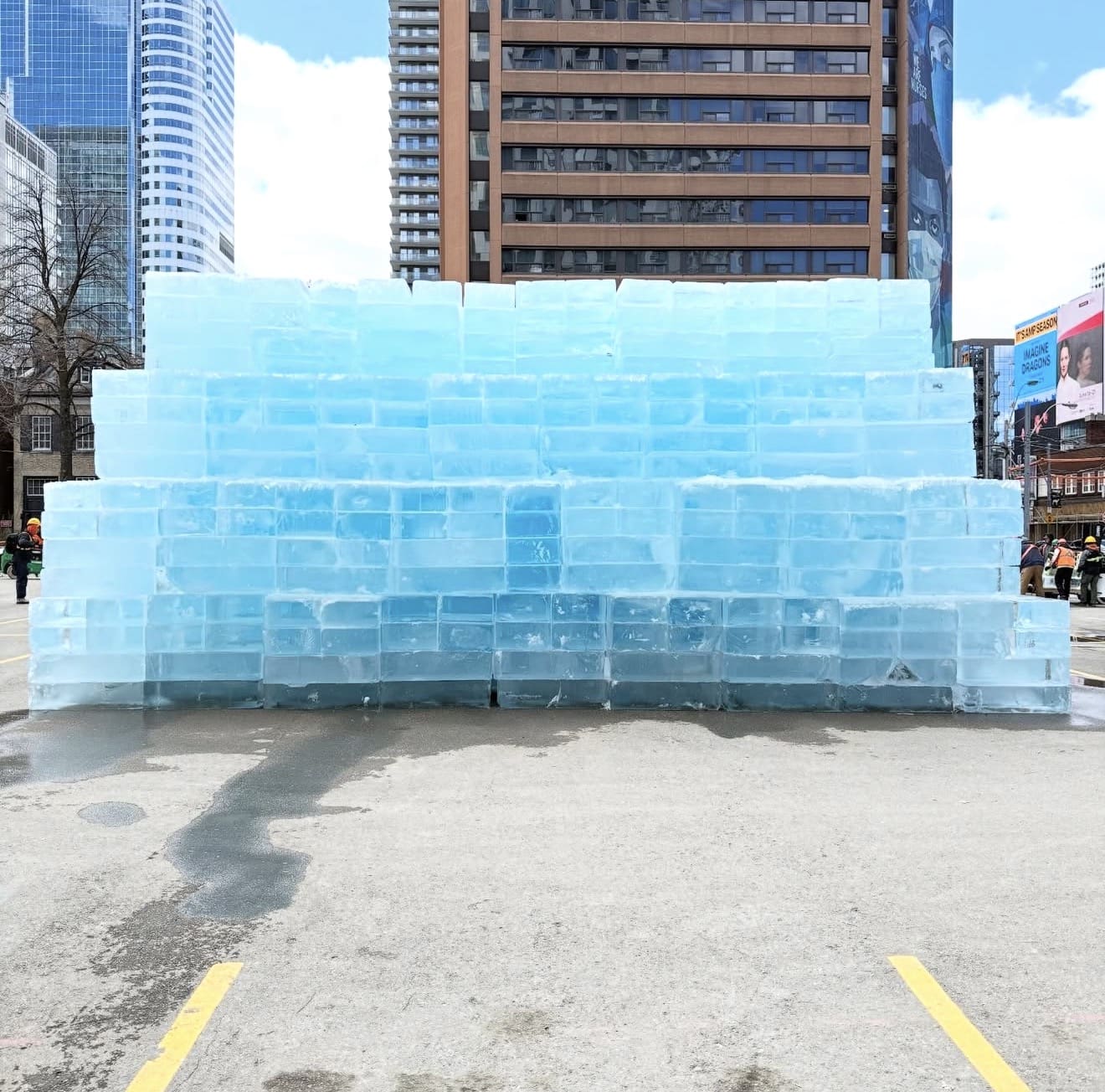 Un immense mur de blocs de glace translucides empilés occupant un stationnement de Toronto, avec les gratte-ciels du centre-ville visibles en arrière-plan.