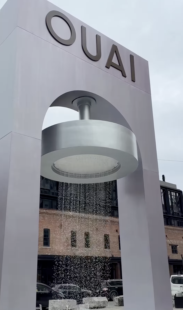 A towering white architectural installation branded "OUAI" with a giant functional shower head raining water down onto a New York City street.