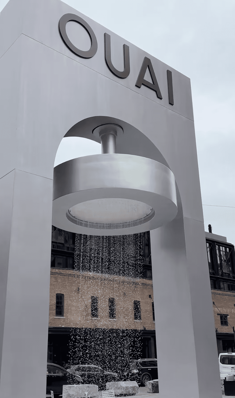 Une imposante installation architecturale blanche de la marque OUAI, avec une gigantesque pomme de douche fonctionnelle faisant tomber de l'eau sur une rue de New York.