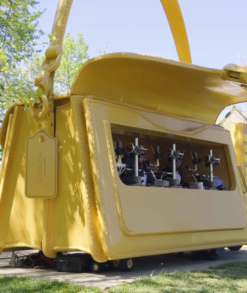 A giant yellow Coach Tabby handbag prop on wheels, large enough to function as a vehicle, with machinery visible through a side window panel.