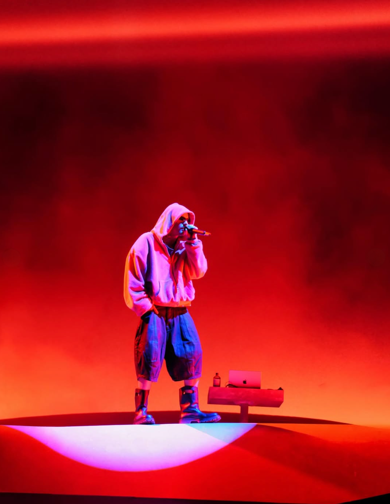 performer in a pink hoodie, baggy shorts, and chunky boots stands alone on a circular lit stage platform, bathed in deep red light, holding a microphone.