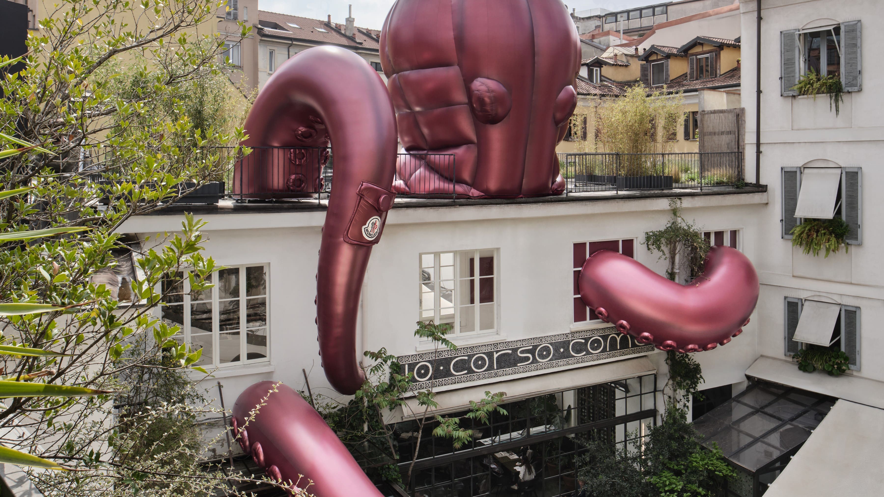 D'immenses tentacules gonflables bordeaux de créatures marines enveloppant la façade du 10 Corso Como à Milan, chacun portant le badge emblématique de Moncler et des détails de poche matelassée.