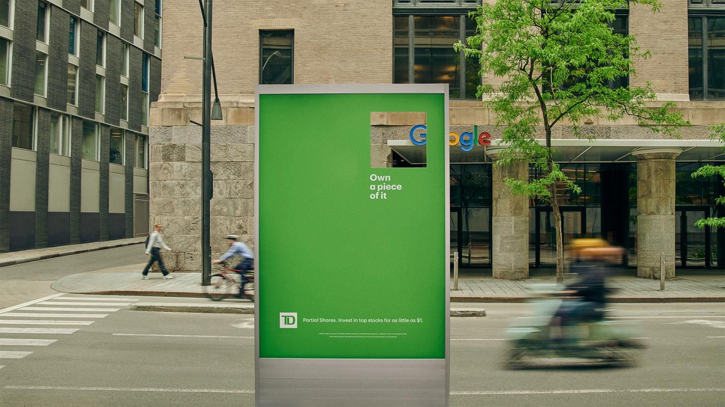 Un panneau d'affichage vert TD Bank avec une petite découpe rectangulaire encadrant le logo Google sur un immeuble en arrière-plan, avec le texte « Own a piece of it » et un slogan sur les actions fractionnées.